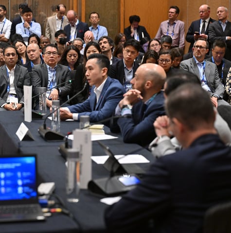 A speaker makes the case at a roundtable, with a full room of delegates listening behind